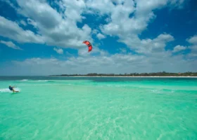 Un atleta fa kitesurf sfrecciando sulle acque turchesi e trasparenti dell'Oceano Indiano davanti alla spiaggia del Medina Palms. Sullo sfondo una striscia di sabbia bianca finissima con vegetazione tropicale e un cielo azzurro parzialmente coperto da nuvole bianche e soffici.