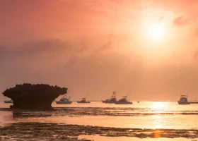 Suggestivo tramonto tropicale nei pressi del Medina Palms Kenya, con il sole che illumina il cielo di calde tonalità arancioni e rosa. La luce si riflette sulla distesa d'acqua a bassa marea, dove galleggiano diverse barche da pesca vicino a un faraglione di roccia scura.