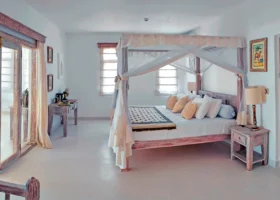 Suite di lusso al Medina Palms Kenya caratterizzata da un grande letto matrimoniale con baldacchino in legno e zanzariera bianca. Arredi in legno decapato stile swahili, pavimento chiaro lucido, porte finestre con infissi rustici che si aprono sulla veranda e pareti bianche decorate con quadri minimali.