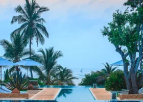 Prospettiva simmetrica di una piscina a sfioro geometrica con camminamenti in legno chiaro al Medina Palms Resort. L'acqua della piscina si fonde visivamente con l'orizzonte dell'oceano dove naviga una dhow, incorniciata da palme e alberi tropicali sotto un cielo sfumato all'alba.