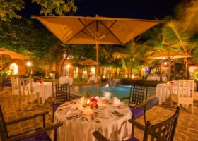 Tavoli allestiti per una cena romantica all'aperto di notte intorno a una piscina illuminata con fontana centrale al Medina Palms. Atmosfera calda creata da lanterne soffuse, ombrelloni aperti, sedie con cuscini viola e palme illuminate sullo sfondo della corte interna del resort.