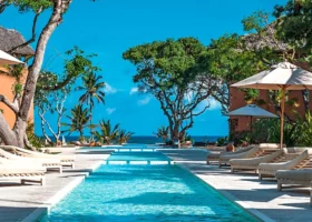 Ampia piscina rettangolare affiancata da terrazze in legno con file di lettini prendisole bianchi e ombrelloni aperti al Medina Palms Watamu. L'architettura del resort color ocra è circondata da alberi e palme che si stagliano contro un cielo blu intenso, con vista sul mare in fondo.