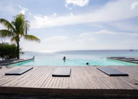Piscina a sfioro sul mare del My Blue Hotel circondata da un solarium in legno con lettini e una palma da cocco sotto un cielo parzialmente nuvoloso