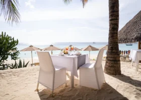 Tavolo per due persone apparecchiato con tovaglia bianca sulla sabbia all'ombra di una struttura in makuti del ristorante del My Blue Hotel