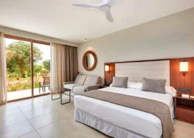 Interno di una moderna camera da letto dell'Hotel Riu Jambo con un letto matrimoniale, un divano grigio e un'ampia finestra affacciata sul giardino tropicale
