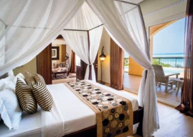 Interno di una lussuosa camera da letto stile villaggio al Riu Palace Zanzibar con letto a baldacchino in legno e veli bianchi, pavimento in parquet chiaro, salottino sullo sfondo e veranda esterna privata con sedia a sdraio affacciata sul mare turchese di Nungwi.
