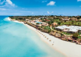 Veduta aerea panoramica della costa del Riu Palace Zanzibar, con la spiaggia di sabbia bianca finissima attrezzata con ombrelloni di paglia, l'oceano Indiano cristallino dalle sfumature azzurre e i giardini tropicali del resort con i blocchi delle camere.