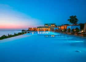 Grande piscina a sfioro del Riu Palace Zanzibar con acqua turchese illuminata, circondata da lettini prendisole con gazebo bianchi e palme, con vista panoramica sull'oceano aperto durante un tramonto tropicale dalle sfumature rosa, arancioni e blu.