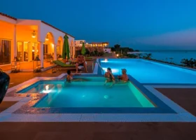 Area relax esterna del Riu Palace Zanzibar di sera con luci calde artificiali della struttura che illuminano una vasca idromassaggio quadrata in primo piano dove si rilassano degli ospiti, accanto alla piscina a sfioro principale affacciata sul mare scuro al tramonto.
