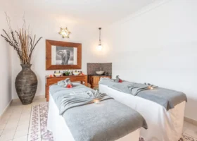 Interno del centro benessere spa del Riu Palace Zanzibar con due lettini da massaggio pronti per i trattamenti, coperti da teli grigi e decorati con fiori rossi, pareti bianche, un grande specchio con cornice in legno massiccio, un quadro con elefante e candele accese.
