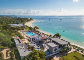 Veduta aerea dal drone del complesso centrale del Riu Palace Zanzibar, che mostra i tetti piani degli edifici moderni, i patii esterni con i tavoli del ristorante, la piscina rettangolare azzurra, i lettini con ombrelloni e il litorale sabbioso con barche ormeggiate in lontananza.
