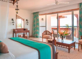 Interno di una spaziosa camera del Royal Zanzibar Beach Resort con un grande letto matrimoniale, tende turchesi e balcone esterno arredato