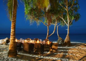 Un lungo tavolo in legno allestito per una cena serale sulla sabbia all'ombra delle palme illuminate al Sultan Sands Island Resort