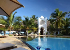 Due cocktail blu in primo piano disposti sul bordo della piscina del Sultan Sands Island Resort di fronte a una struttura bianca in stile zanzibarino