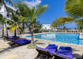 La grande piscina all'aperto del Watamu Beach Resort circondata da una pavimentazione in pietra chiara, lettini prendisole in legno con materassini e asciugamani viola, piante di palme e la struttura del resort sullo sfondo.