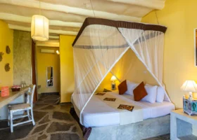 Interno della camera superior del Watamu Beach Resort arredata in stile afro-chic con un grande letto matrimoniale dotato di zanzariera bianca a baldacchino, pareti gialle, pavimento in pietra scura e scrivania in legno rustico.
