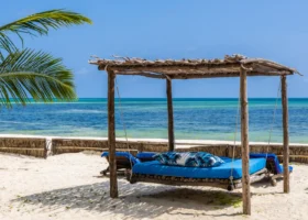 Un tradizionale letto a baldacchino in legno grezzo con materasso blu e cuscino a motivi tropicali posizionato sulla sabbia bianca della spiaggia privata del Watamu Beach Resort, con vista sul mare del Kenya.