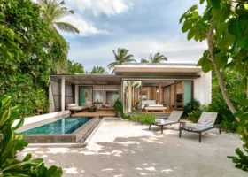 Esterno di una beach villa dal design contemporaneo con ampie vetrate aperte su camera e living, deck in legno con piscina privata a sfioro e lettini sulla sabbia bianca, incorniciata da fitta vegetazione tropicale e palme da cocco. Alila Kothaifaru Maldives, Isola di Kothaifaru, Atollo di Raa