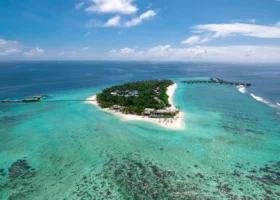 Veduta aerea dell'intera isola di Kothaifaru coperta di fitta vegetazione, circondata dalla spiaggia bianca, dalla barriera corallina affiorante e dalla lunga fila di ville overwater che si protende nella laguna turchese dell'Oceano Indiano. Alila Kothaifaru Maldives, Atollo di Raa, Maldive.