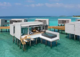 Veduta delle ville overwater dal design architettonico contemporaneo in pietra chiara, sospese su palafitte sopra la laguna turchese cristallina, ciascuna con deck privato, piscina a sfioro e scaletta di accesso diretto all'acqua. Alila Kothaifaru Maldives, Kothaifaru, Atollo di Raa, Oceano Indiano.