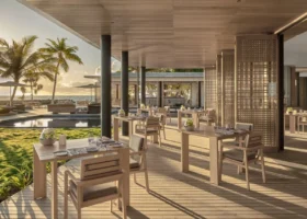 Ristorante open-air dal design contemporaneo in legno chiaro con tavoli e sedie apparecchiati, paraventi a griglia e ampie aperture affacciate su prato, piscina e oceano nella luce dorata del tramonto tra le palme. Alila Kothaifaru Maldives, Kothaifaru, Atollo di Raa, Maldive.