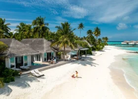 Veduta aerea delle beach villa dal tetto in strame allineate lungo la spiaggia di sabbia bianca, con piscine private a sfioro e deck in legno; una coppia rilassata in terrazza e due bambini che giocano sulla sabbia, palme da cocco e la laguna turchese con le ville overwater sullo sfondo. Isola di Maagau, Atollo di Dhaalu, Maldive.