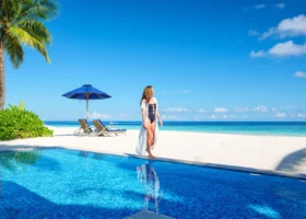 Una donna in costume nero e lungo copricostume bianco cammina sul bordo di una piscina a sfioro affacciata sulla spiaggia; accanto un ombrellone blu e lettini sulla sabbia bianca, con l'oceano che passa dal turchese al blu profondo all'orizzonte. Baglioni Maldives, Maagau, Atollo di Dhaalu.