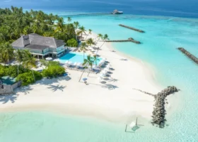 Veduta aerea della zona piscina e spiaggia del resort, con l'edificio principale dal tetto in strame, la piscina a sfioro, lettini e ombrelloni allineati sulla sabbia bianca e i frangiflutti in roccia che si protendono nella laguna turchese. Isola di Maagau, Atollo di Dhaalu, Maldive.
