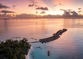 Veduta aerea dell'isola al tramonto, con il cielo arancio e dorato e la lunga fila di ville overwater che curva sulla laguna dai riflessi del sole; in primo piano il palmeto e la spiaggia bianca. Maagau, Atollo di Dhaalu, Maldive, Oceano Indiano.