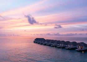 Lunga fila di ville overwater dal tetto in strame allineate sulla laguna al tramonto, con le luci calde accese e il cielo rosa e lilla che si riflette sull'acqua calma. Baglioni Maldives, Isola di Maagau, Atollo di Dhaalu.