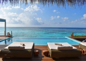 Terrazza in legno di una water villa con piscina privata a sfioro e due daybed in rattan affacciati sull'Oceano Indiano; il tetto in strame incornicia la vista aperta sulla barriera corallina e sul cielo azzurro. Baglioni Maldives, Maagau, Atollo di Dhaalu.