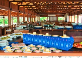 Lungo buffet di dolci e dessert con runner in tessuto ikat blu, torte, crostatine e roll di pan di spagna disposti su piattini quadrati, vassoio di frutta fresca e ampia struttura in legno con travi a vista e vetrate, al The Barefoot Eco Hotel sull'isola di Hanimaadhoo, Atollo di Haa Dhaalu, Maldive.