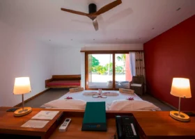 Interno di una camera Beach con parete d'accento rossa, ventilatore a pale in legno, due lampade da comodino accese e letto matrimoniale decorato con petali e fiori di frangipani, porta scorrevole affacciata sul giardino e sulla spiaggia, al The Barefoot Eco Hotel, isola di Hanimaadhoo, Atollo di Haa Dhaalu, Maldive.