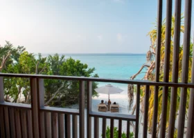 Vista sull'Oceano Indiano turchese dal balcone in legno di una camera Ocean View, con ombrellone bianco e due lettini sulla spiaggia di sabbia bianca incorniciata da vegetazione tropicale e palme, al The Barefoot Eco Hotel, isola di Hanimaadhoo, Atollo di Haa Dhaalu, Maldive.