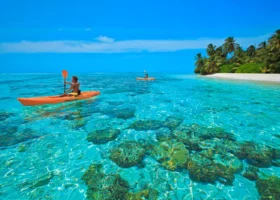 Due persone pagaiano su kayak arancioni nella laguna turchese e cristallina sopra un fondale corallino, lungo la costa di un'isola orlata di palme e sabbia bianca, al The Barefoot Eco Hotel, isola di Hanimaadhoo, Atollo di Haa Dhaalu, Maldive, Oceano Indiano.