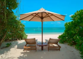 Due lettini in legno e ombrellone beige su sabbia bianca finissima, incorniciati da fitta vegetazione tropicale, affacciati sul mare turchese con un dhoni maldiviano all'ancora, al The Barefoot Eco Hotel, isola di Hanimaadhoo, Atollo di Haa Dhaalu, Maldive.