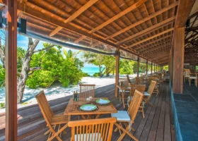 Terrazza ristorante coperta con tetto e travi in legno, tavoli e sedie in teak apparecchiati con piatti verdi e tovaglioli, affacciata sulla spiaggia di sabbia bianca e sul mare turchese oltre la vegetazione, al The Barefoot Eco Hotel, isola di Hanimaadhoo, Atollo di Haa Dhaalu, Maldive.
