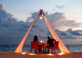 Coppia a una cena romantica privata al crepuscolo sotto un baldacchino di tessuto bianco illuminato da lanterne, sulla spiaggia di sabbia bianca di Brennia Kottefaru affacciata sull'Oceano Indiano, Atollo di Raa, Maldive.