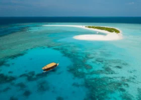 Lingua di sabbia bianca deserta con un dhoni maldiviano tradizionale ancorato nella laguna turchese e cristallina di Brennia Kottefaru, Atollo di Raa, Maldive, con la barriera corallina chiaramente visibile sotto la superficie dell'acqua.