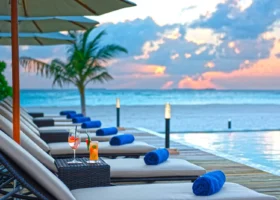 Lettini prendisole con teli arrotolati blu e ombrelloni allineati lungo la piscina a sfioro di Brennia Kottefaru al tramonto, con due cocktail colorati su un tavolino e una palma affacciata sull'Oceano Indiano, Atollo di Raa, Maldive.