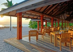 Ristorante all'aperto di Brennia Kottefaru con tavoli e sedie in legno di teak sotto una tettoia con travi a vista, direttamente sulla spiaggia di sabbia bianca al tramonto, affacciato sull'Oceano Indiano, Atollo di Raa, Maldive.