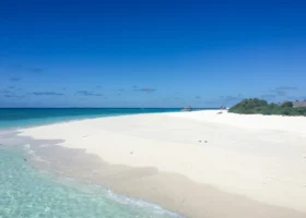 Ampia spiaggia di sabbia bianca finissima e deserta di Brennia Kottefaru che degrada nella laguna turchese e cristallina dell'Atollo di Raa, alle Maldive, con vegetazione tropicale sulla destra e l'Oceano Indiano all'orizzonte.
