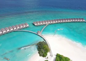 Vista aerea delle ville overwater di Brennia Kottefaru allineate su pontili in legno sopra la laguna turchese dell'Atollo di Raa, alle Maldive, con la spiaggia di sabbia bianca e l'Oceano Indiano blu profondo sullo sfondo.