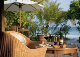 Aperitivo romantico su una terrazza in legno di Coco Palm Dhuni Kolhu, Atollo di Baa, Maldive: due calici di vino rosso, bottiglia, tagliere di formaggi e ciotole di olive su un tavolino rotondo in teak, poltrona in vimini intrecciato in primo piano sfocata, ombrellone bianco, palme e vegetazione tropicale illuminate dalla luce calda del tramonto e spiaggia con oceano sullo sfondo in profondità di campo.