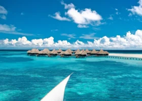 Dhoni maldiviano in legno con vela bianca spiegata naviga sulla laguna turchese di Coco Palm Dhuni Kolhu, isola di Dhuni Kolhu, Atollo di Baa, Maldive; sullo sfondo la fila di ville overwater con tetto in makuti collegate da un pontile, cielo blu intenso con nubi cumuliformi e fondali corallini visibili in trasparenza nell'Oceano Indiano.