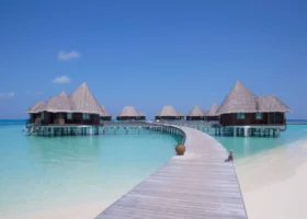 Pontile in legno chiaro che parte dalla spiaggia di sabbia bianca e curva sulla laguna verso le ville overwater di Coco Palm Dhuni Kolhu, isola di Dhuni Kolhu, Atollo di Baa, Maldive: bungalow su palafitte bianche con alti tetti conici in makuti e facciate in legno scuro, anfora in terracotta appoggiata sul camminamento, acqua trasparente su fondale sabbioso e cielo azzurro limpido sull'Oceano Indiano.