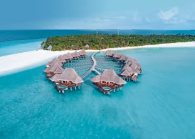 Ripresa aerea in pieno giorno del gruppo di ville overwater di Coco Palm Dhuni Kolhu, Atollo di Baa, Maldive: bungalow su palafitte con tetto conico in makuti disposti a ventaglio e collegati da pontili in legno, piscine idromassaggio private azzurre sulle terrazze, laguna turchese brillante su fondale sabbioso, punta di sabbia bianca e isola coperta di palme sullo sfondo con l'Oceano Indiano all'orizzonte.