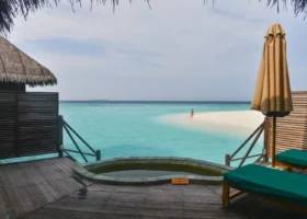 Terrazza in legno di una water villa di Coco Palm Dhuni Kolhu, isola di Dhuni Kolhu, Atollo di Baa, Maldive: piscina idromassaggio privata a filo del deck con targa della profondità, lettini con cuscini verde smeraldo e ombrellone chiuso color sabbia, paravento in doghe di legno scuro e tetto in makuti sulla sinistra, laguna turchese trasparente e lingua di sabbia bianca raggiungibile a piedi dove una persona sosta in acqua bassa, con l'Oceano Indiano blu profondo all'orizzonte.