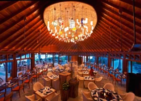Interno serale di un ristorante dall'alto tetto a volta in legno con una scenografica installazione di lampade colorate in vetro sospese al centro, tavoli apparecchiati con tovaglie bianche e candele e ampie vetrate affacciate sulla spiaggia. Constance Halaveli, Halaveli, Atollo di Ari Nord.