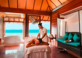 Sala massaggi della spa overwater dal soffitto in legno caldo, con una terapista che pratica un trattamento su un lettino, vasca freestanding e daybed dai cuscini turchesi affacciati attraverso le ampie aperture sulla laguna turchese. Constance Halaveli, Isola di Halaveli, Atollo di Ari Nord.